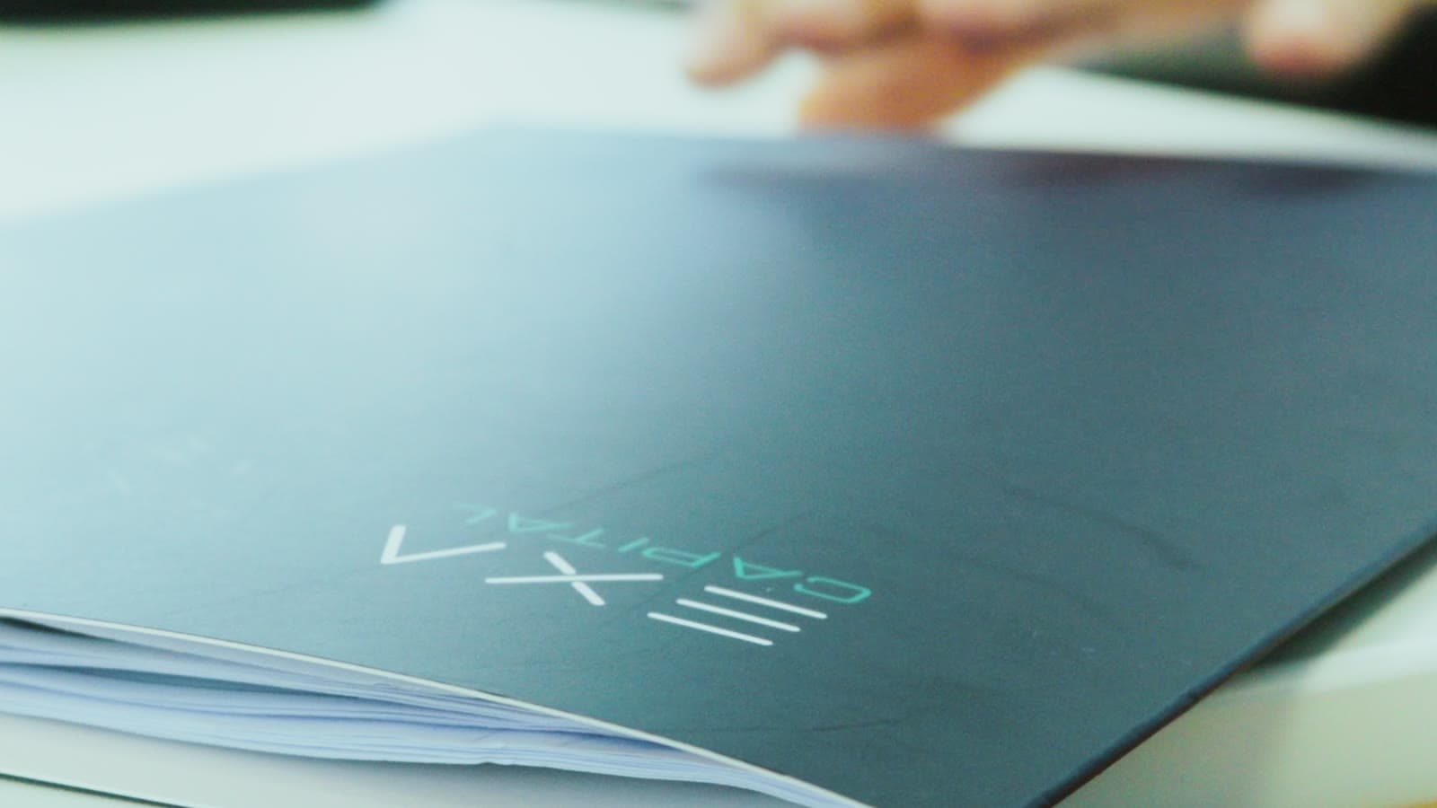 Frame from the Exa Capital film: a hand opening a navy branded folder on a desk.
