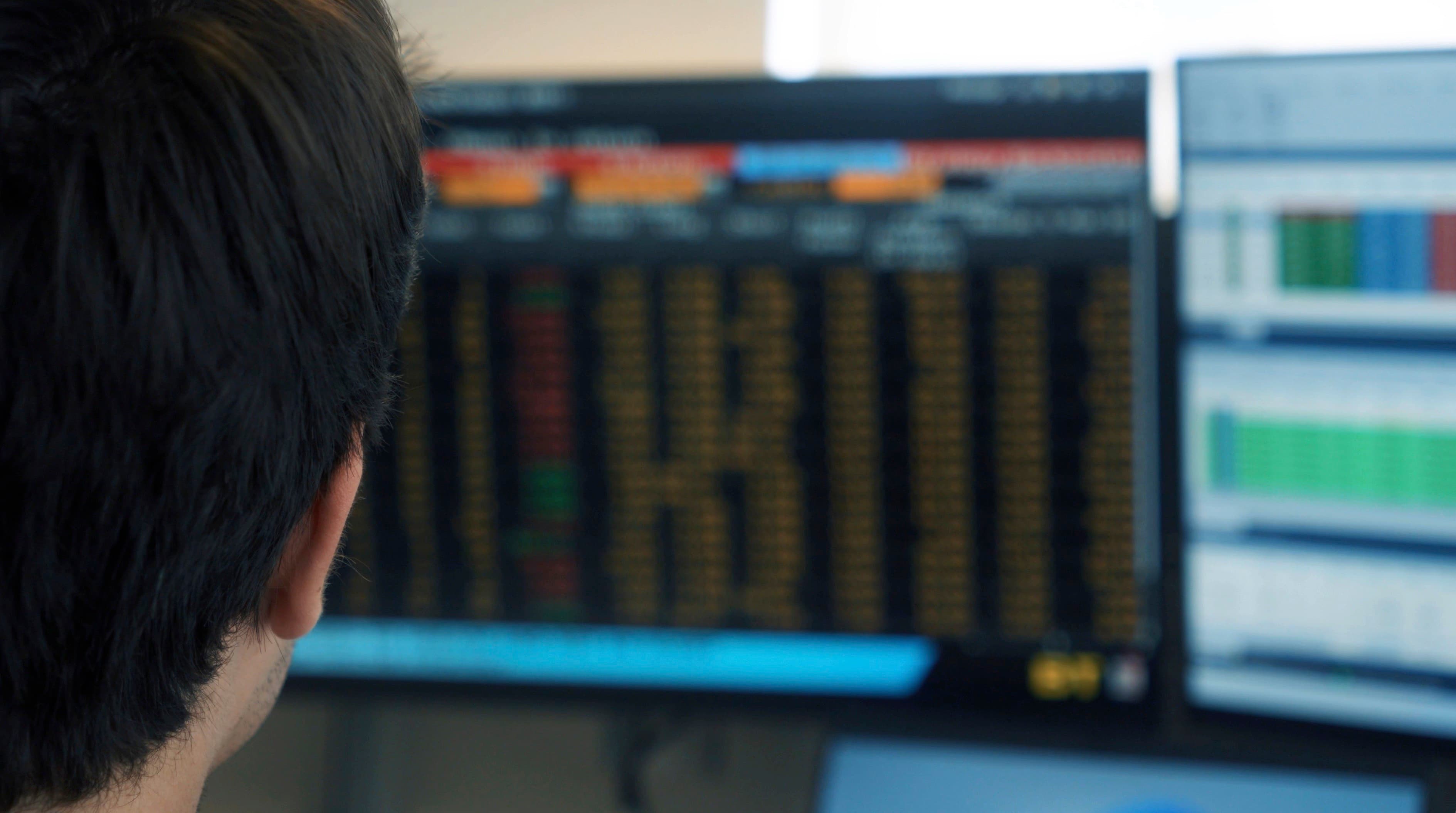 Over a shoulder: a monitor filled with columns of live figures, a second screen beside it.