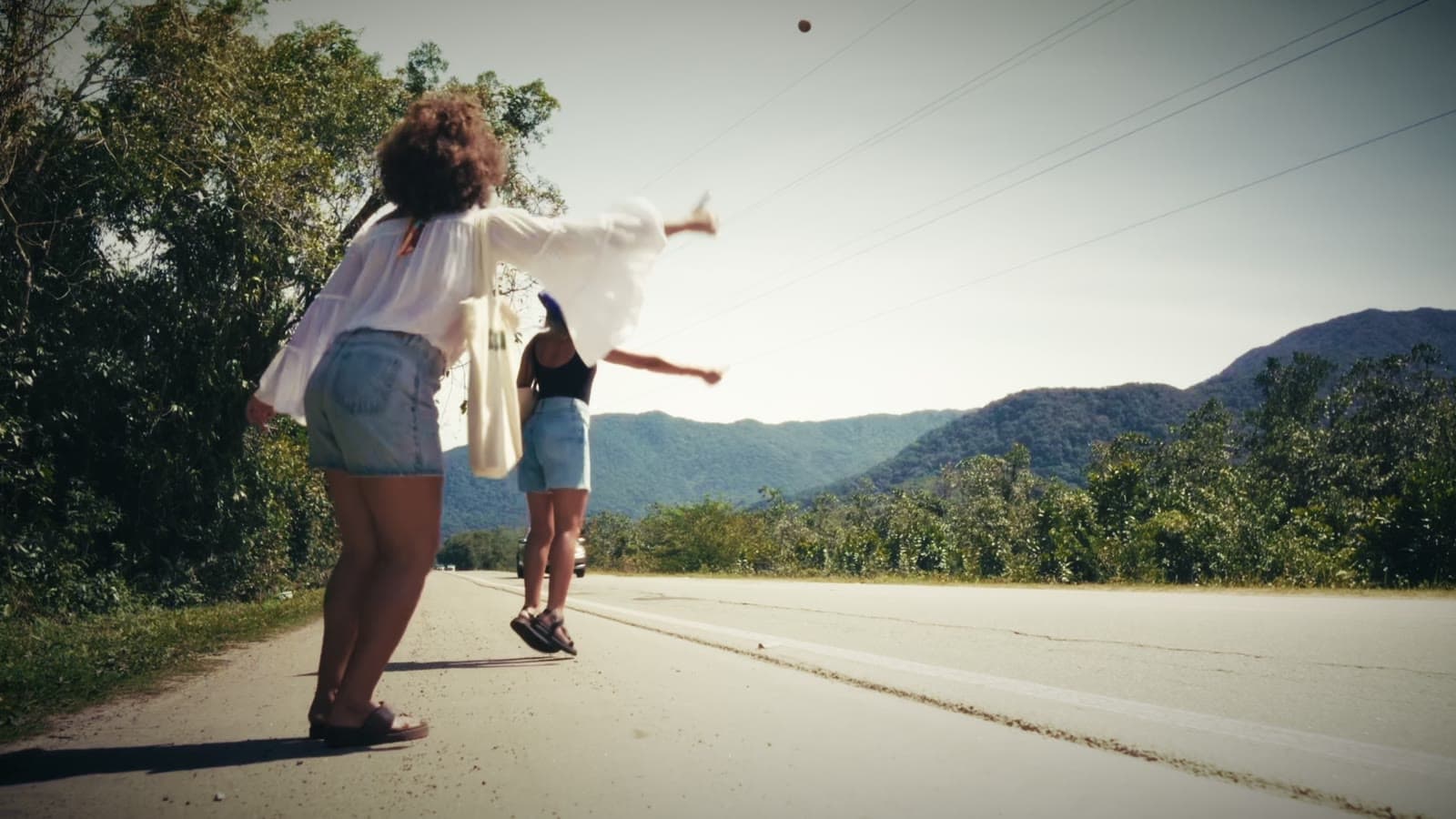 Frame from the Rota 55 film: two travellers on the shoulder of the coastal highway, forested hills behind.